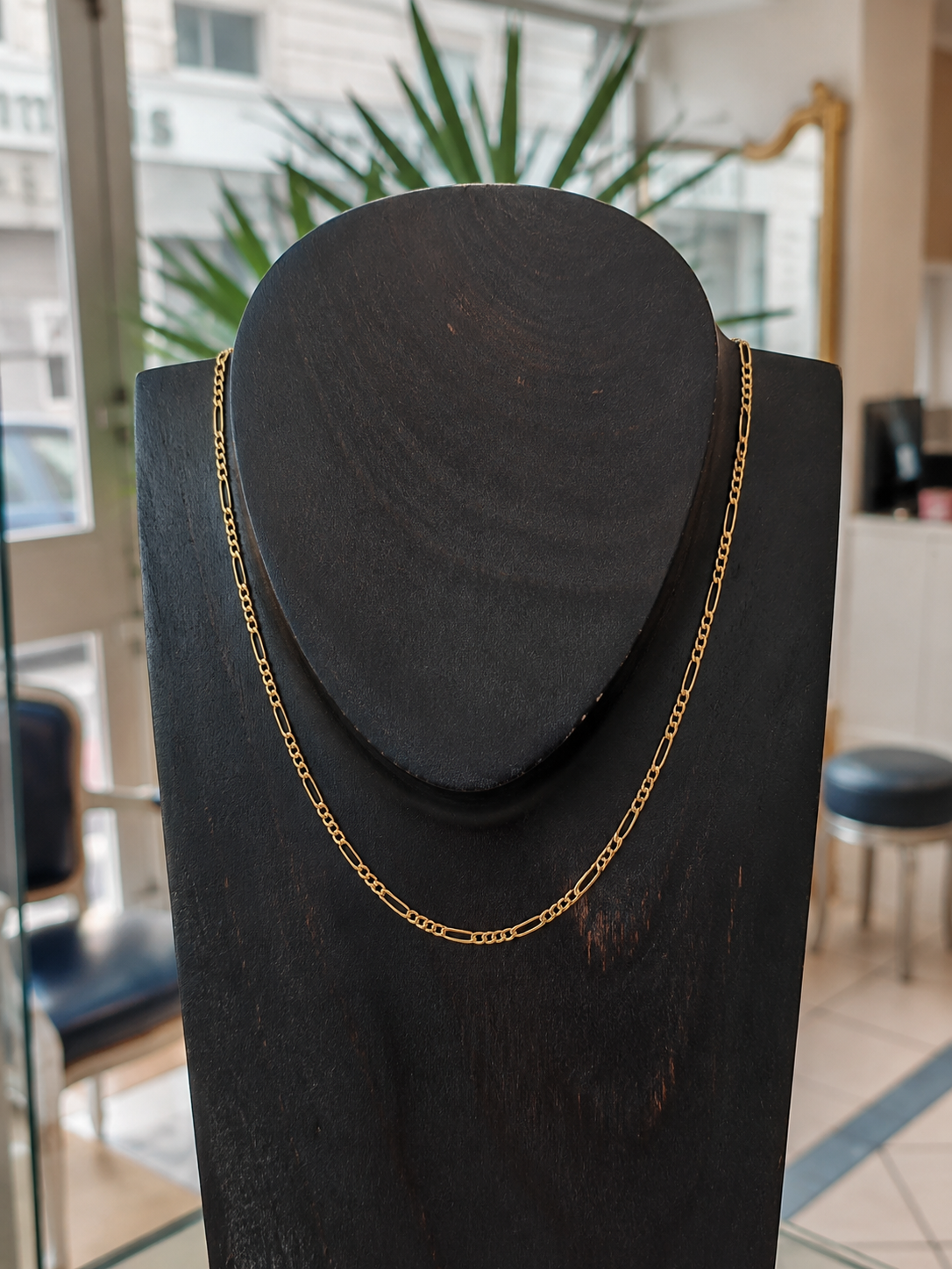 Chaine en or cheval figaro 1/3 en or jaune 18 carats sur un bustier en bois noir en premier plan, en second plan on y voit l'accueil d'une belle bijouterie