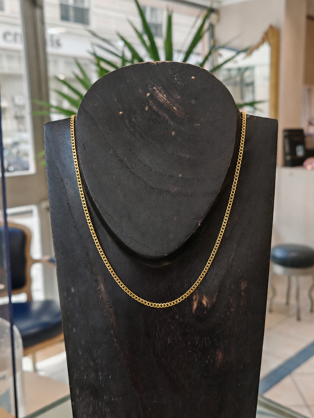 Chaine en or gourmette en or jaune 18 carats sur un bustier en bois noir en premier plan, en second plan on y voit l'accueil d'une belle bijouterie