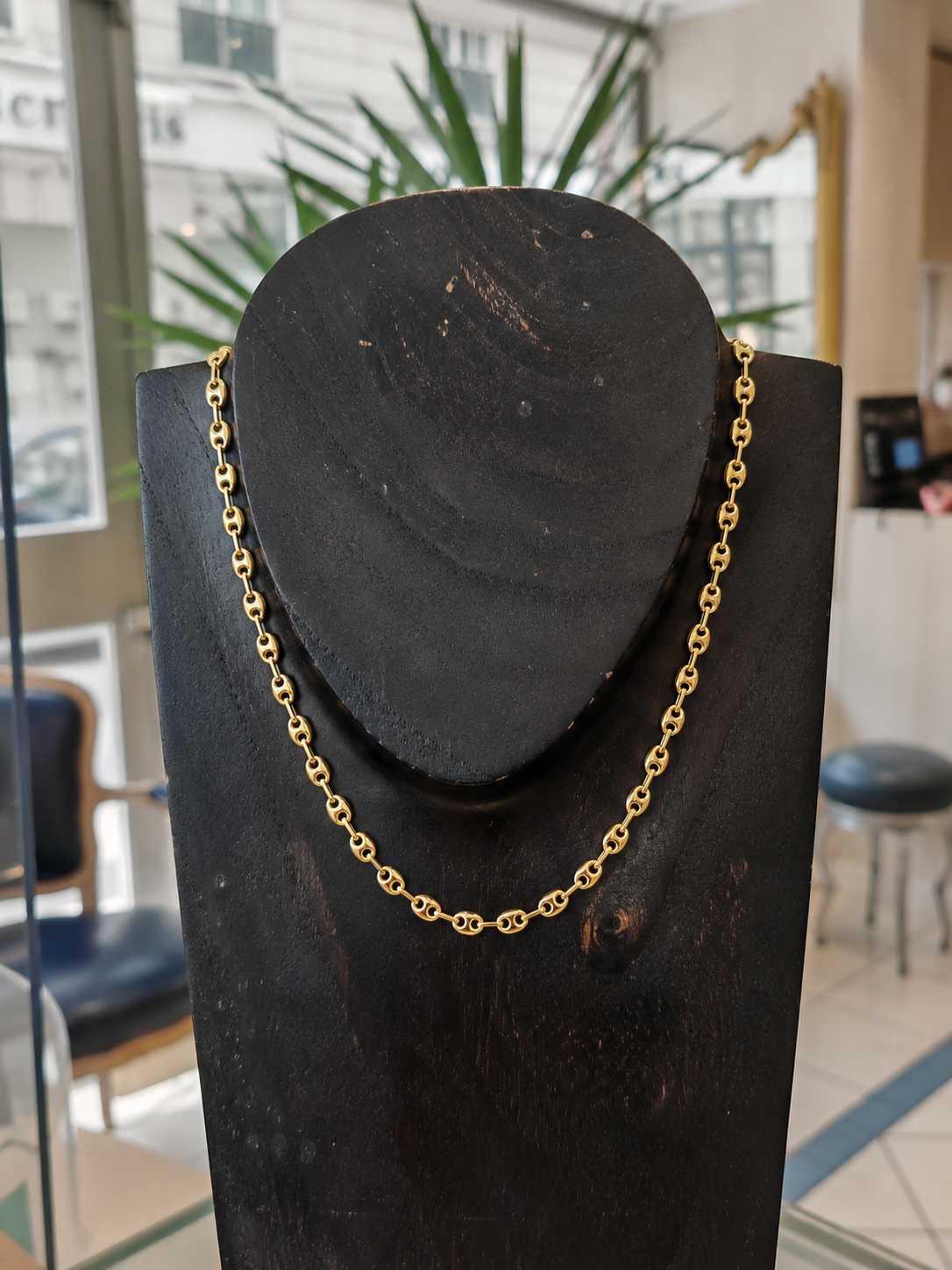 Chaine en or grain de café en or jaune 18 carats sur un bustier en bois noir en premier plan, en second plan on y voit l'accueil d'une belle bijouterie