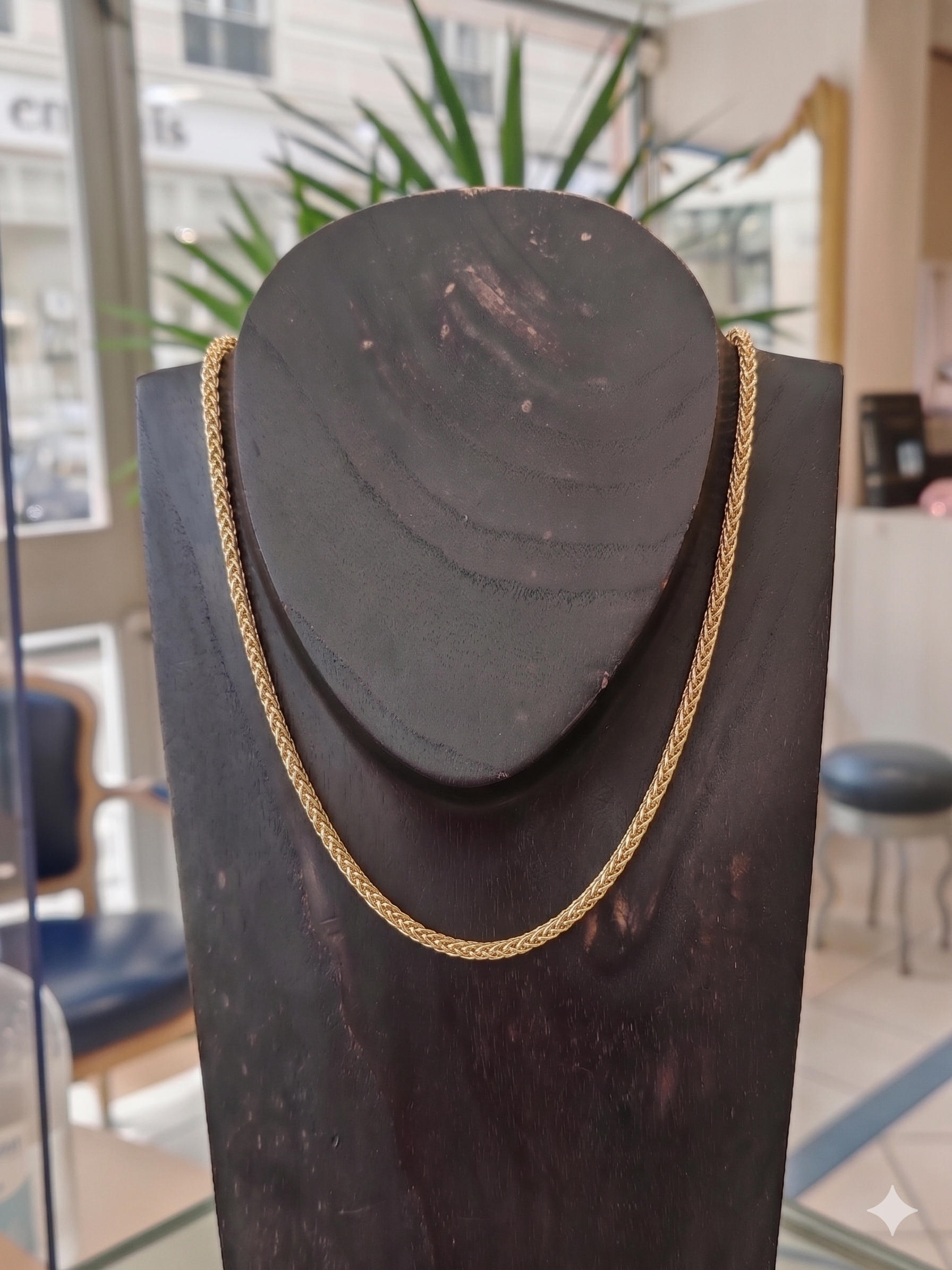Chaine en or palmier en or jaune 18 carats sur un bustier en bois noir en premier plan, en second plan on y voit l'accueil d'une belle bijouterie